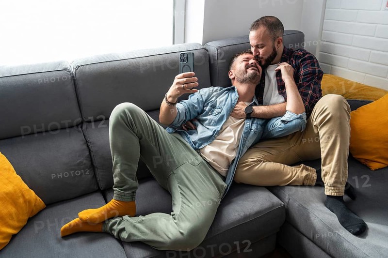 A gay couple relaxes on a sofa, taking a selfie. One partner lies on the other's lap, showcasing a moment of affection and comfort in a bright living room
