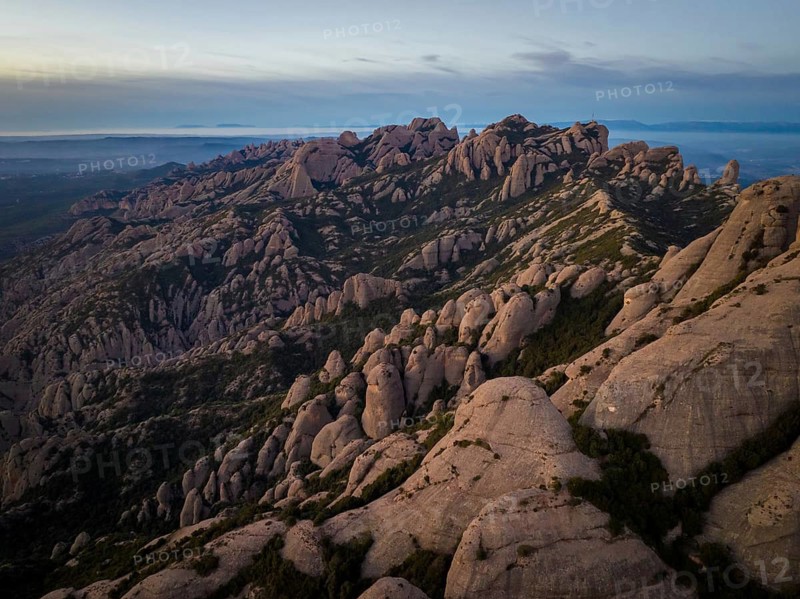 Aerial view of dramatic rock formations in Montserrat, Catalonia, at sunset. The rugged landscape reveals reddish stone peaks contrasted against a serene sky