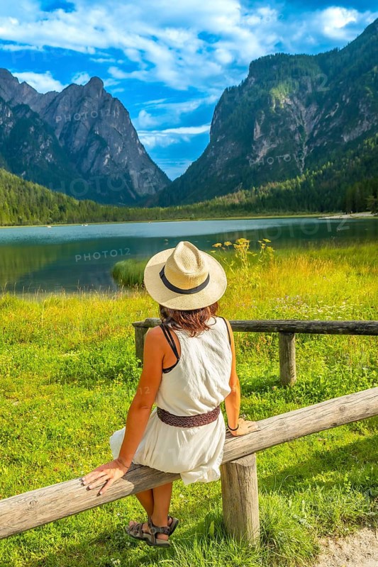 Woman wearing summer hat sitting on a wooden fence, admiring lake dobbiaco and the dolomites, italy