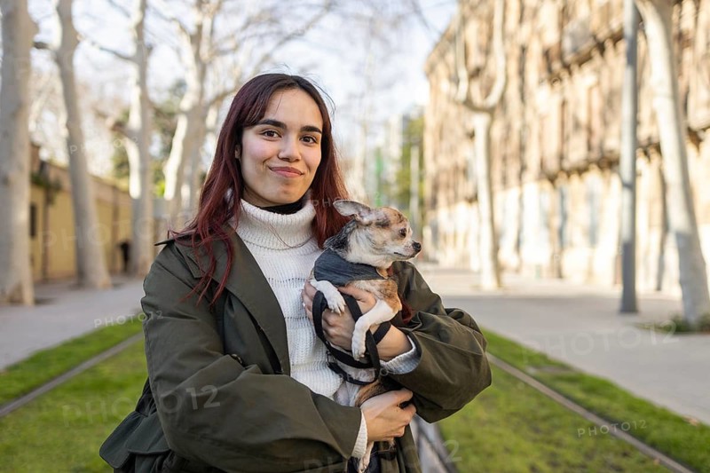 A woman with long dark hair smiles while holding her chihuahua, enjoying a sunny day in a tree lined street wearing a cozy white sweater and green jacket