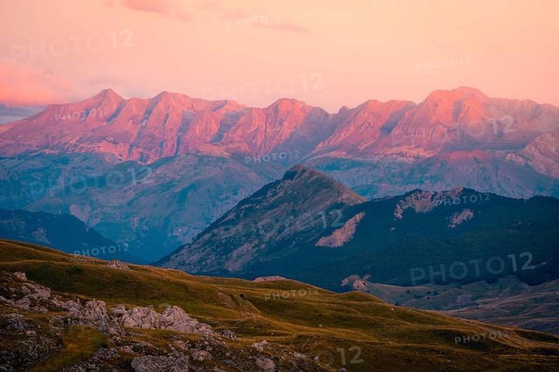 A stunning mountain range illuminated by the soft glow of sunset, featuring vibrant pink and orange hues in summer. Rolling hills in the foreground add depth to this serene landscape in the Pyrenees