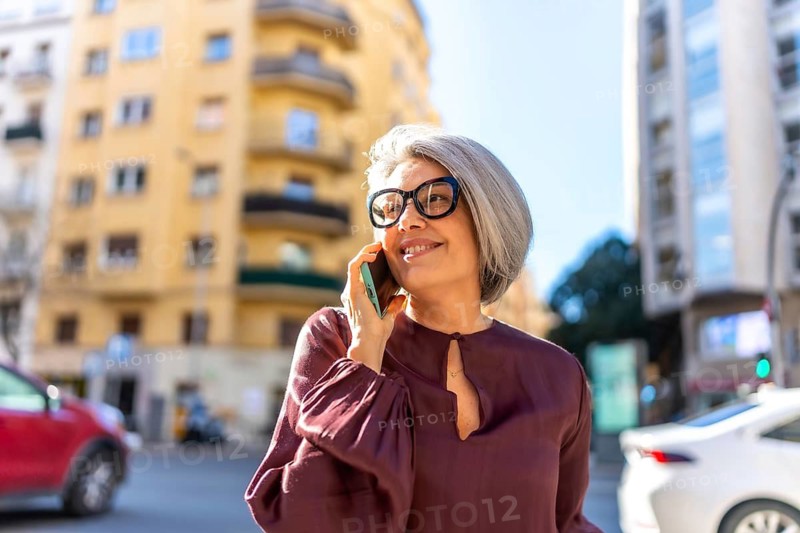 Stylish gray haired woman with glasses smiling while talking on her smartphone outdoors in a sunny city street, confident and modern, enjoying a casual urban lifestyle