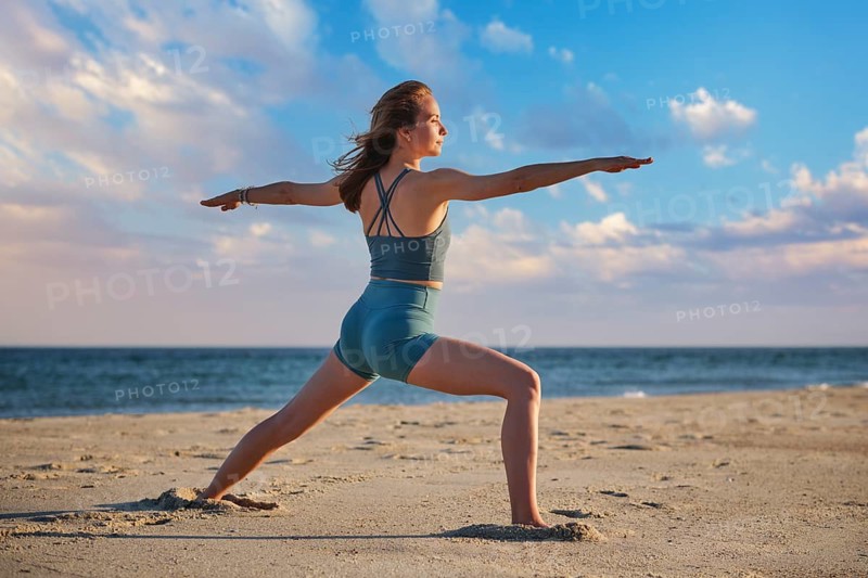 A woman performs Virabhadrasana 2, or Warrior 2 pose, on a beach during sunset, enjoying the peaceful atmosphere and views