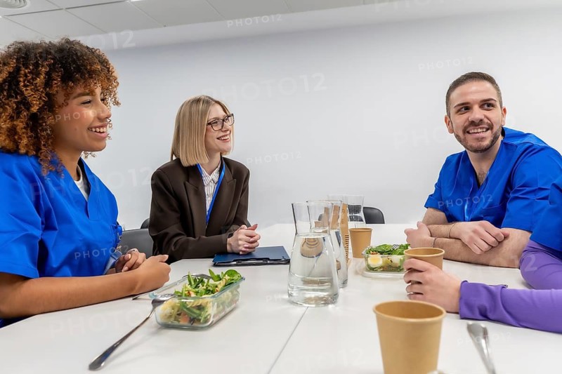 Diverse team of healthcare workers and a businesswoman enjoying a cheerful lunch break at a clinic or hospital, sharing a healthy meal and conversation