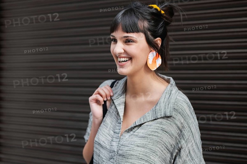 A cheerful young woman smiles while against a dark backdrop. She wears a casual gray shirt, vibrant orange earrings, and carries a black bag over her shoulder