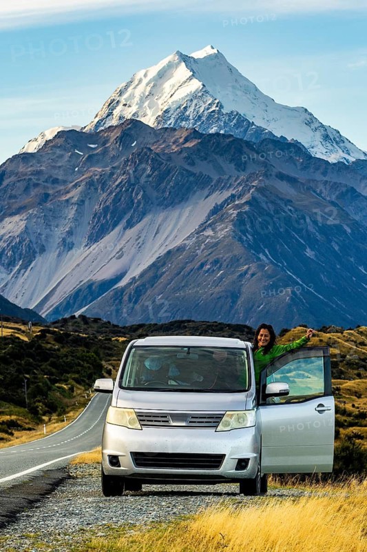 A woman stands beside her parked car, enjoying the stunning autumn view of Mount Cook in New Zealand. The majestic mountain landscape highlights the serene beauty of the season