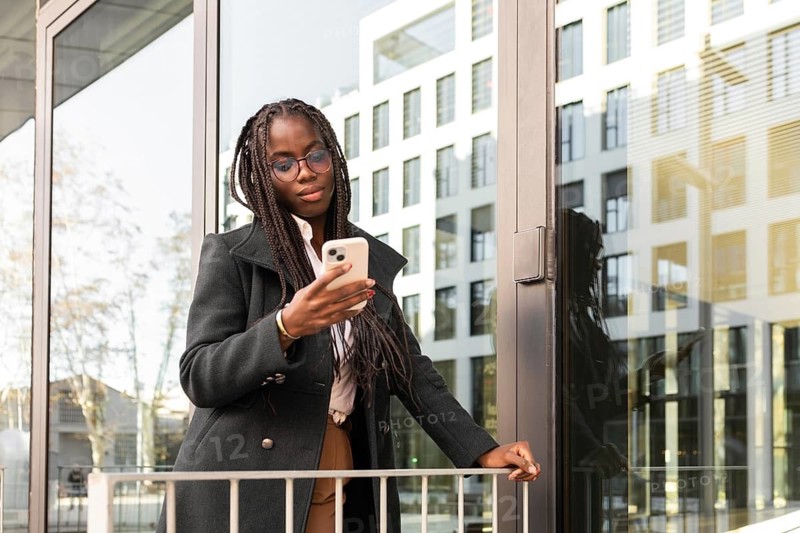 African American businesswoman with braids using a smartphone during a break outside a modern office building