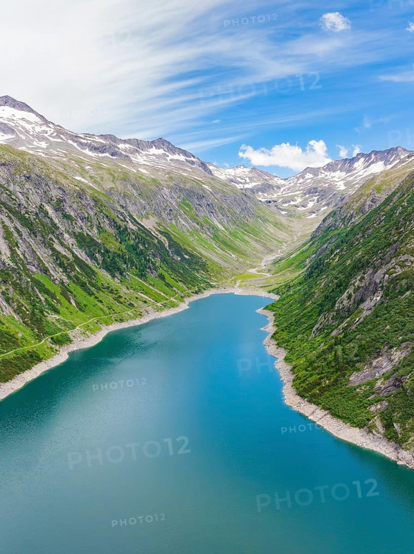 Mountain landscape with an elongated lake in the centre, surrounded by green hills and snow-capped mountains under a blue sky, Klein Tibet, Zillertal, Austria