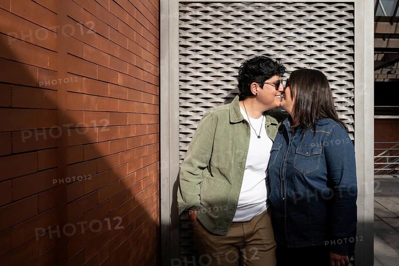 A joyful lesbian couple shares a tender moment outdoors against a brick wall. Both women are casually dressed, expressing love and connection in a public setting