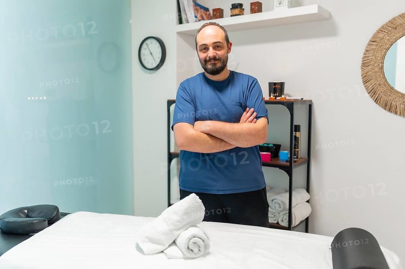 Massage therapist with crossed arms standing in a spa or treatment room, looking at the camera with a friendly expression, ready to provide relaxation and therapeutic care with fresh towels on a bed