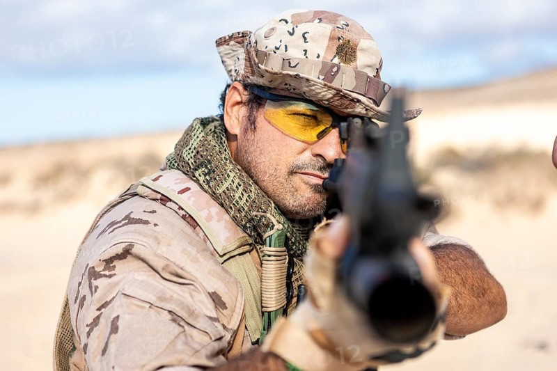 A soldier dressed in desert camouflage aims a rifle with precision and focus. He wears protective eye wear and a tactical helmet, exuding concentration and readiness