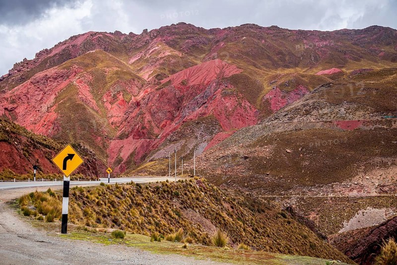 A stunning view of the Peruvian Andes, featuring vibrant red mountain slopes and a winding road with a yellow curve sign. The dramatic sky enhances the scenery