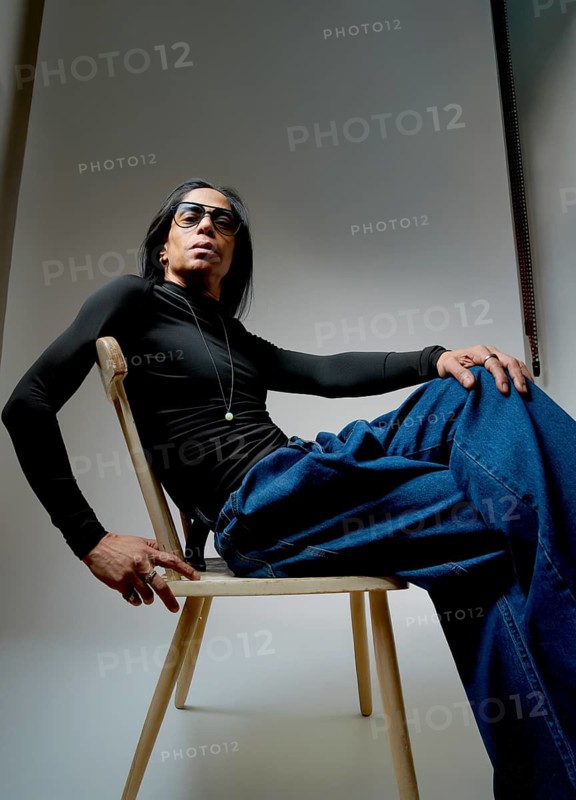 A portrait of a stylish Latin man sitting on a wooden chair, wearing a sleek black long-sleeve shirt and blue jeans. His confident pose exudes casual elegance