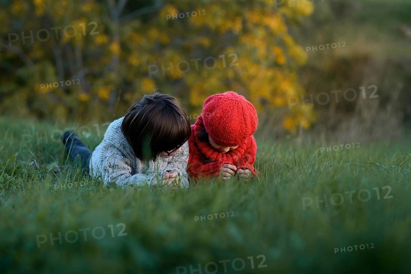 Two siblings, a boy and a girl, are playing on the grass in a park surrounded by autumn foliage. The girl in a red knit hat and the boy in a gray sweater are intently examining something on the ground amidst vibrant fall colors