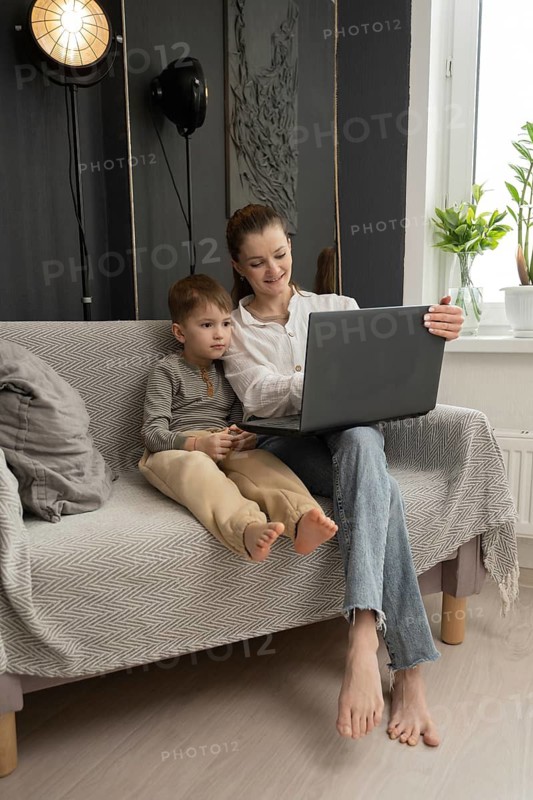 A mother and her son share a bonding moment on a cozy sofa, engaged with a laptop. The scene reflects warmth, technology, and family connection at home