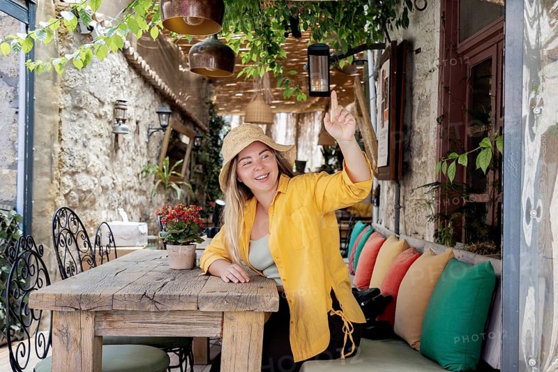 A young, cheerful woman in a yellow shirt sits at a cafe table, wearing a straw hat. She is smiling brightly and gesturing as she seems ready to order
