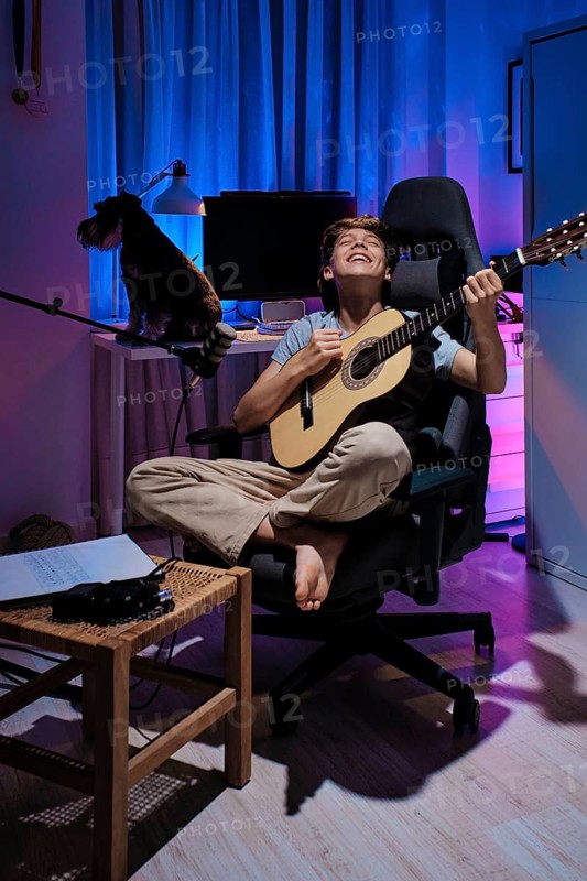 A young guitarist joyfully plays acoustic guitar in a warmly lit room. Their dog sits nearby. The setting features vibrant blue and pink lighting, creating a relaxed atmosphere