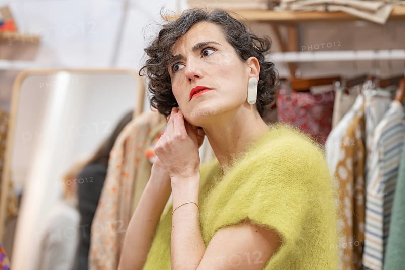A woman in a cozy clothes shop adjusts a bright earring. She wears a soft green sweater, with clothing hanging in the background, creating a warm, stylish scene