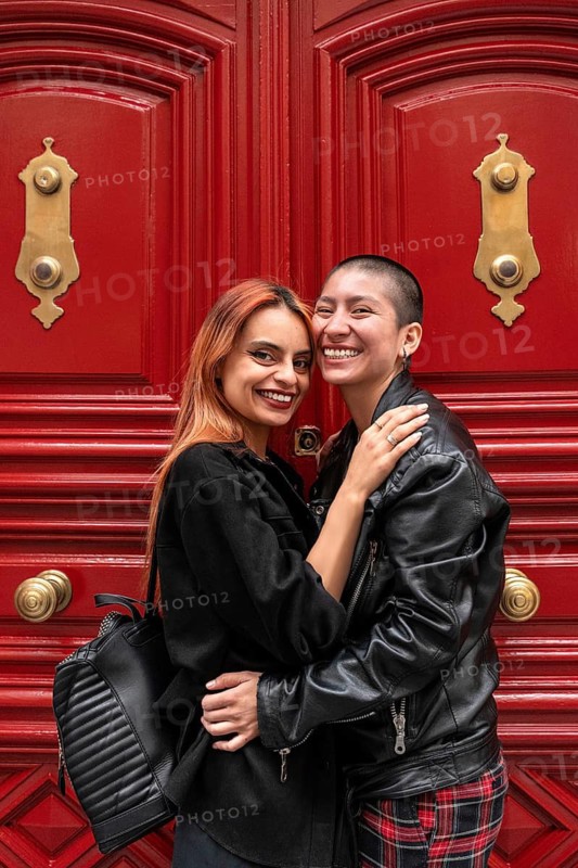 A joyful lesbian couple embraces lovingly in front of striking red doors with golden details. The scene is vibrant, romantic, and highlights genuine affection and happiness