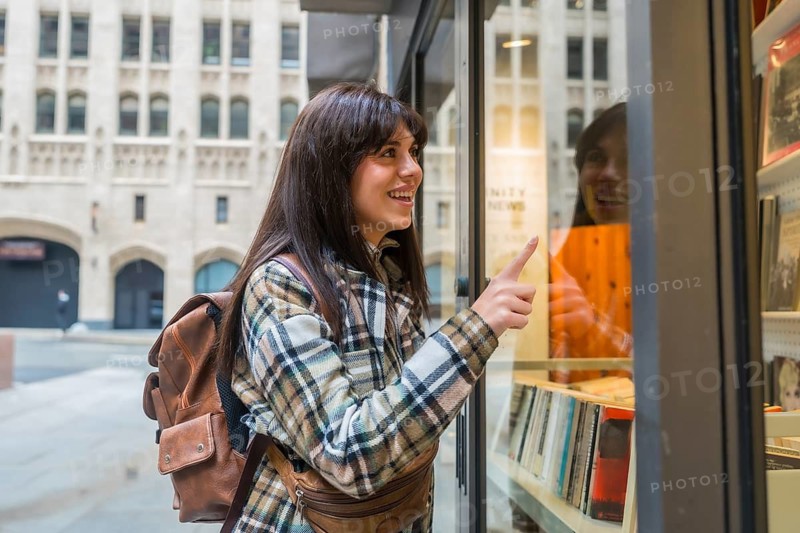 Young woman with a backpack and plaid jacket window shopping, enthusiastically pointing at items in a bookstore display, exploring a vibrant urban street in san francisco