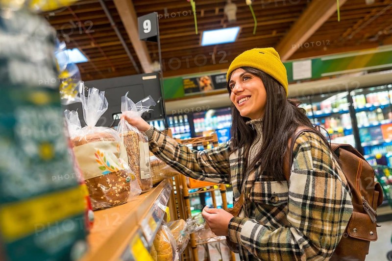 Smiling woman wearing a yellow beanie and plaid shirt selecting fresh healthy bread from a grocery store bakery shelf, enjoying the shopping experience for wholesome food