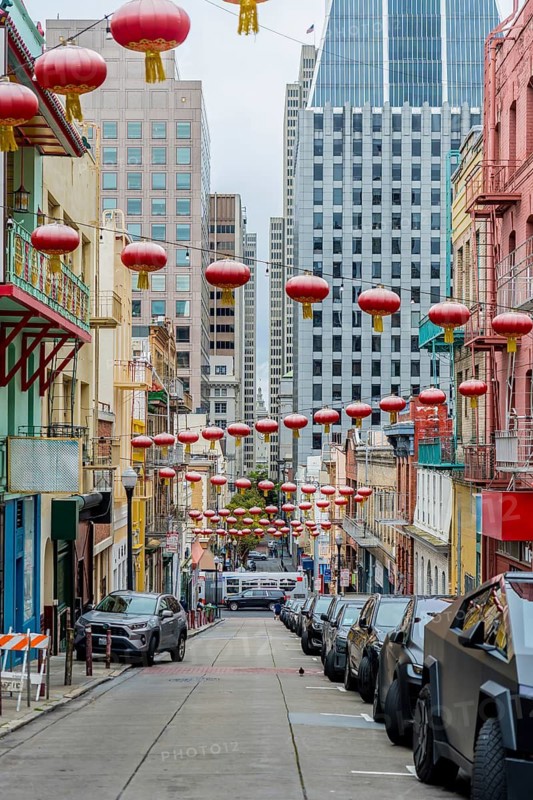 San francisco chinatown street with red lanterns strung above a steep, colorful historic block, parked cars and modern skyscrapers rising in the cloudy background