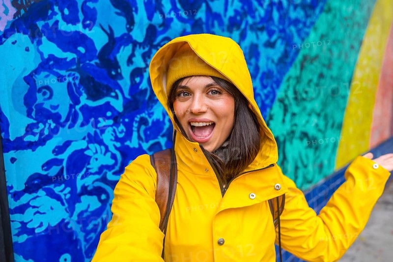 Young woman traveler wearing a yellow raincoat and beanie smiling while taking a selfie on a vibrant urban street, with colorful wall art and murals in the background