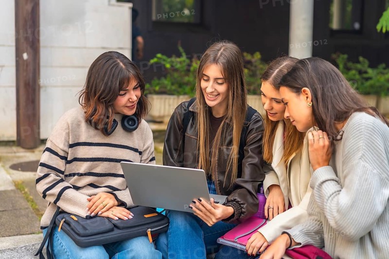 Group of happy female university friends collaborating on a project, learning and sharing knowledge while looking at a laptop computer outdoors on campus