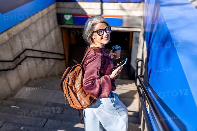 Stylish mature woman with grey hair ascending subway stairs, holding a disposable coffee cup and smartphone, looking over her shoulder and smiling confidently at the camera