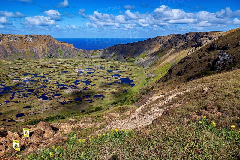 View of a wide crater surrounded by rocks, with the coast and ocean in the background, the Ranu Kau volcano on Easter Island in the Pacific Ocean