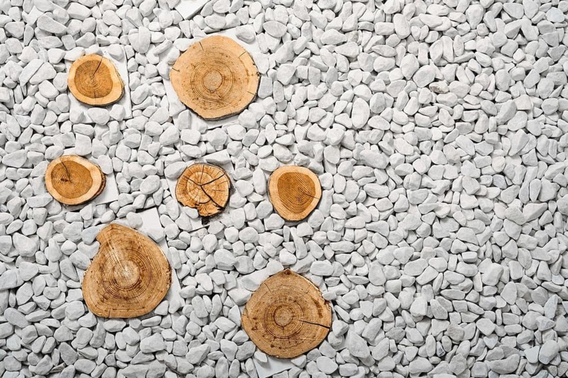 Wooden slices are stuck into the ground and surrounded by small white stones. Simple design suitable for landscaping or decor. The focus is on natural materials used in an outdoor space