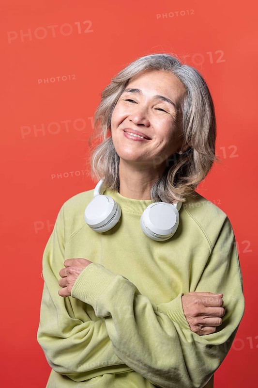 Asian Chinese senior woman wearing a light-colored sweatshirt and headphones, smiling warmly against a vibrant red background, embodying joy and relaxation