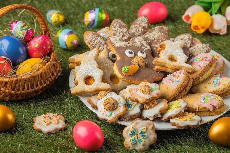A plate with various Easter cookies and a basket full of colorful Easter eggs, decorated with tulip blossoms on grass