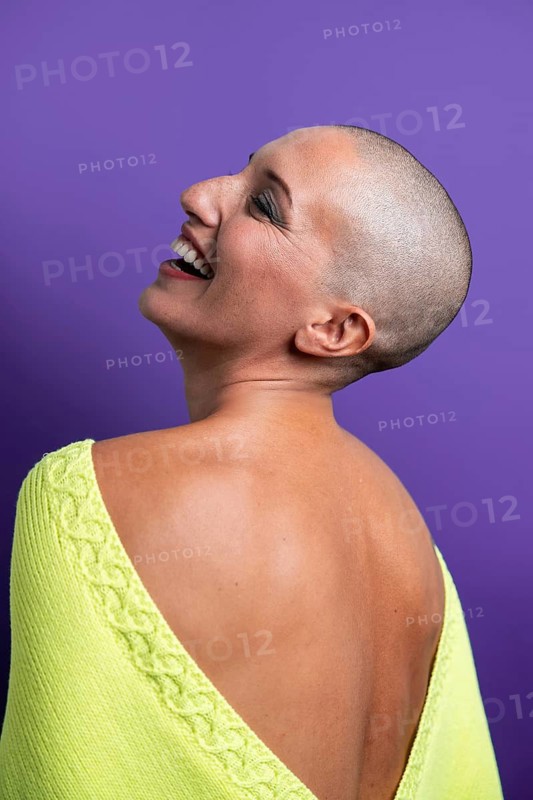 Middle-aged woman with a shaved head wearing a yellow top, smiling and looking up against a purple background, embodying confidence and joy against a vibrant backdrop