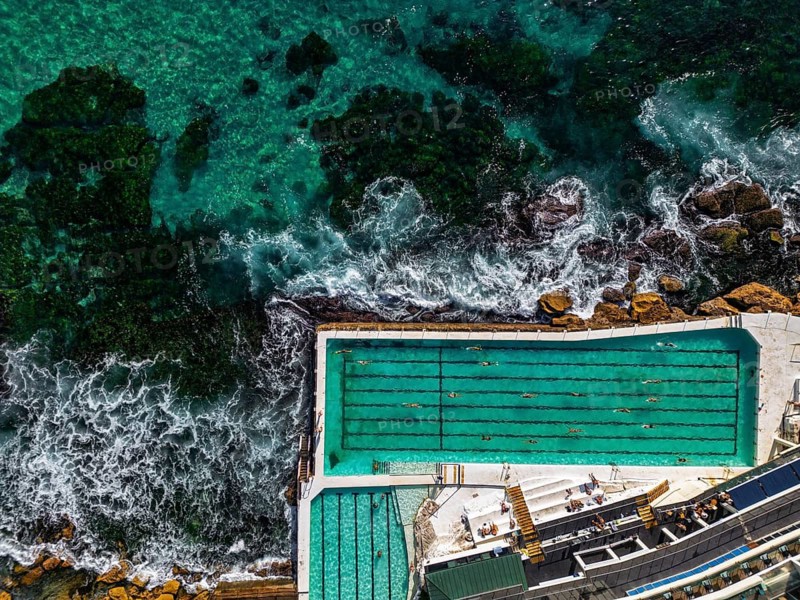 Stunning aerial shot of a seaside pool next to vibrant turquoise ocean, with swimmers enjoying the serene water while waves crash along rocky shore, blending nature and leisure in Sidney, Australia
