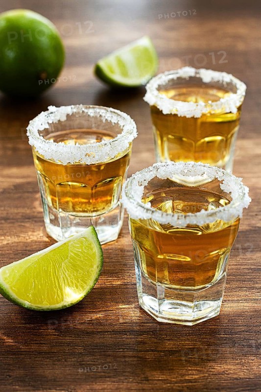 Three glasses with tequila and salt on the rims are arranged on a wooden table. Each glass has a slice of lime on top. A lime sits in the background under natural light
