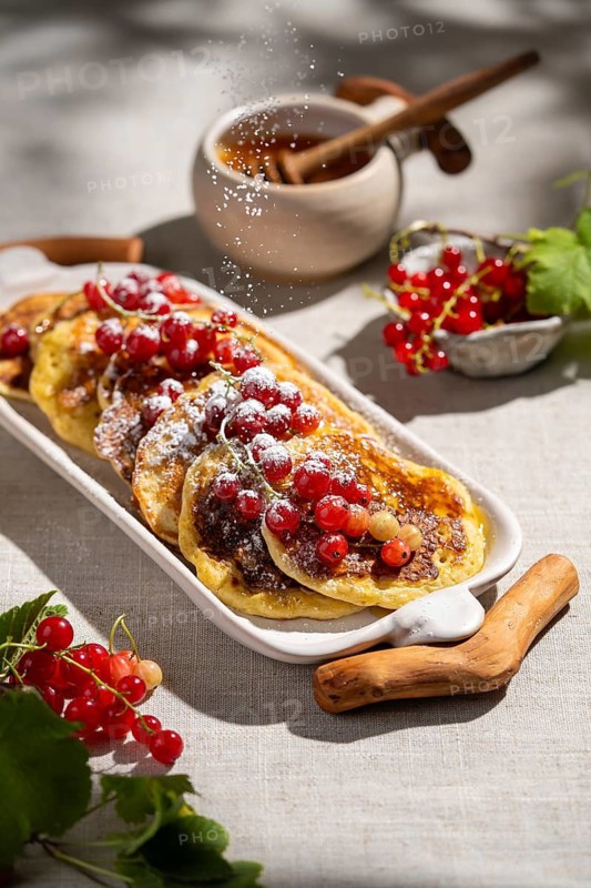 From above, fluffy pancakes are topped with vibrant red currant and dusted with powdered sugar on a white ceramic plate. Background is a light beige fabric
