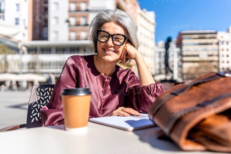Grey haired woman at an outdoor cafe smiling at camera, enjoying sunny city day with coffee, notebook and bag while working remotely or planning her next project