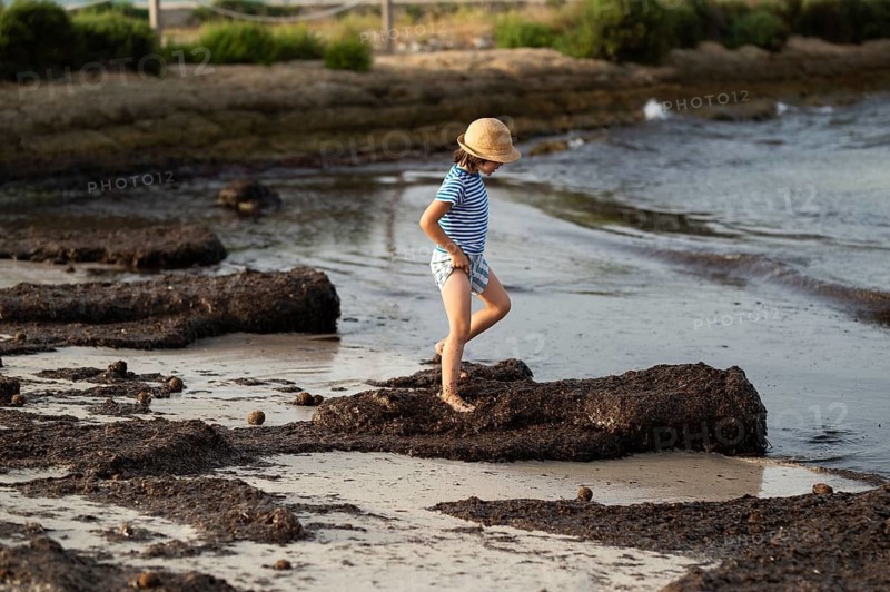 Side view of an unrecognizable young boy wearing a straw hat and striped shirt, carefully stepping on rocks at a beach