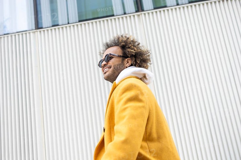 A confident afro man in a bright yellow coat and sunglasses smiles warmly, expressing positivity and style against a modern urban backdrop with vertical lines