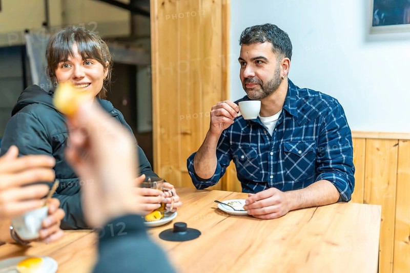 Friends sharing coffee and tea around a warm wooden table in a cozy urban cafe, smiling and chatting in a relaxed, candid social gathering of connection and everyday joy