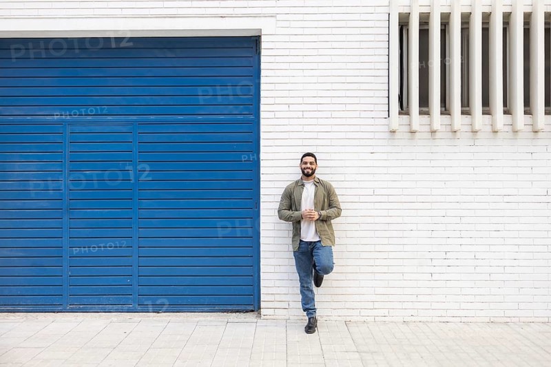 A man stands casually against a white brick wall next to a blue garage door in the city. His relaxed posture and confident smile create a welcoming urban vibe on a sunny day