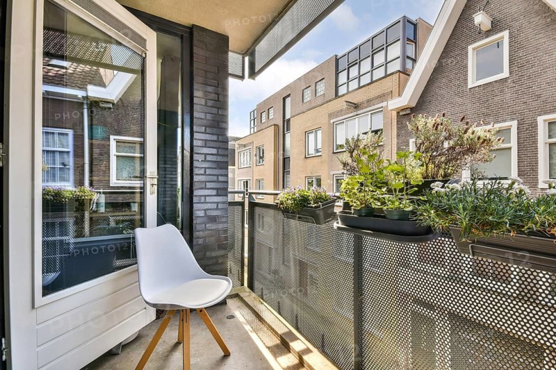 A charming urban balcony features a modern white chair and lush potted plants against a backdrop of city buildings, providing a serene outdoor escape in a bustling environment