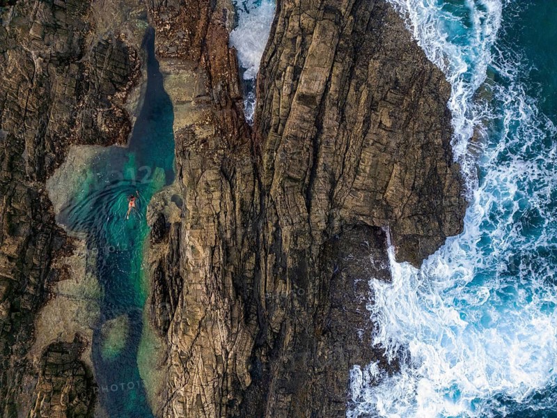 Top view of natural rock pools in Tindaya, Fuerteventura, featuring a woman leisurely swimming in clear blue waters, surrounded by rugged cliffs and waves crashing nearby