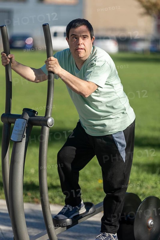 A man with Down syndrome uses outdoor fitness equipment at a park, engaging in physical activity. His focused expression highlights independence, health, and inclusion in an active lifestyle