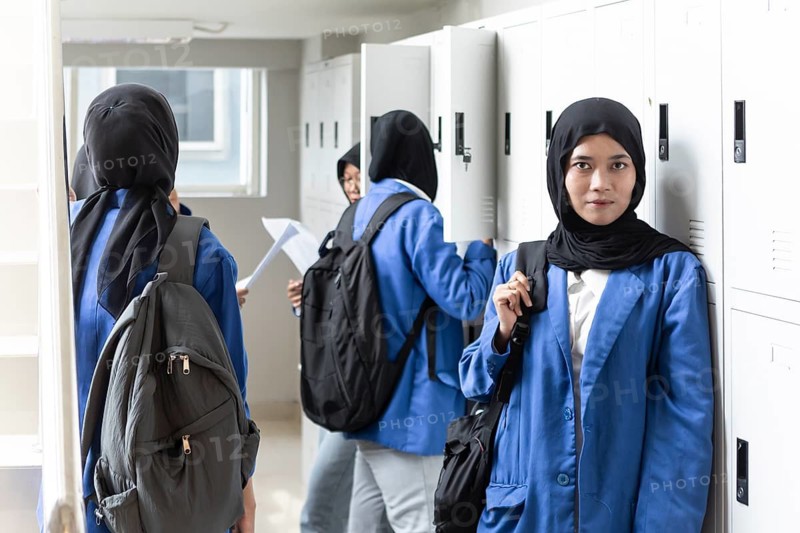 Asian Muslim female students in blue uniforms gather near lockers, preparing for a new school day with backpacks and textbooks, symbolizing education and cultural diversity