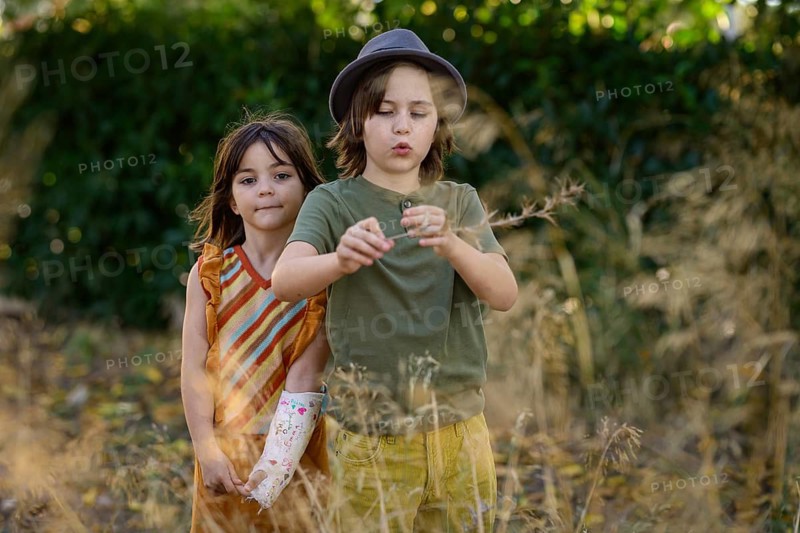 Two children are joyfully playing in a sunlit field. One is wearing a hat and is curious about a plant, while the other watches with an artistic cast on their arm