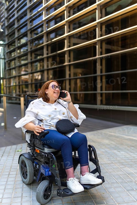 Smiling businesswoman in wheelchair making a phone call in front of a modern office building, enjoying a moment of her working day
