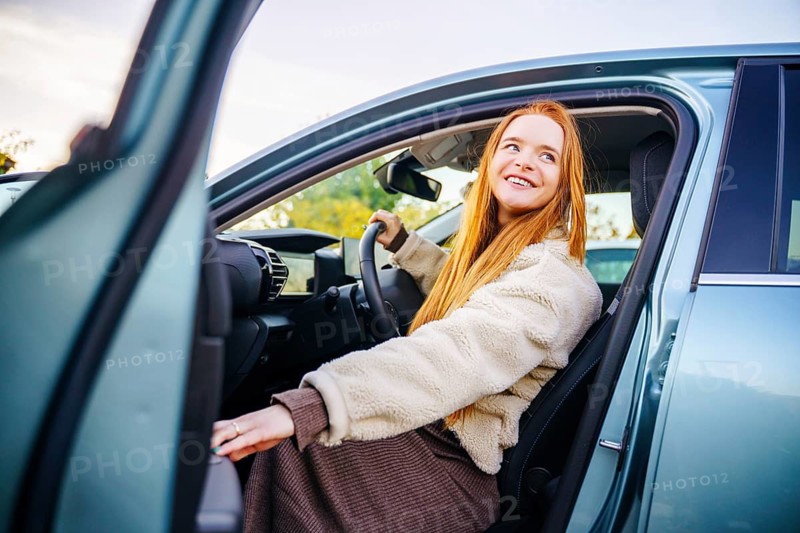 A beautiful pregnant woman with vibrant red hair steps out of a car, smiling brightly. She wears a cozy, warm outfit against a scenic autumn backdrop, radiating happiness and serenity