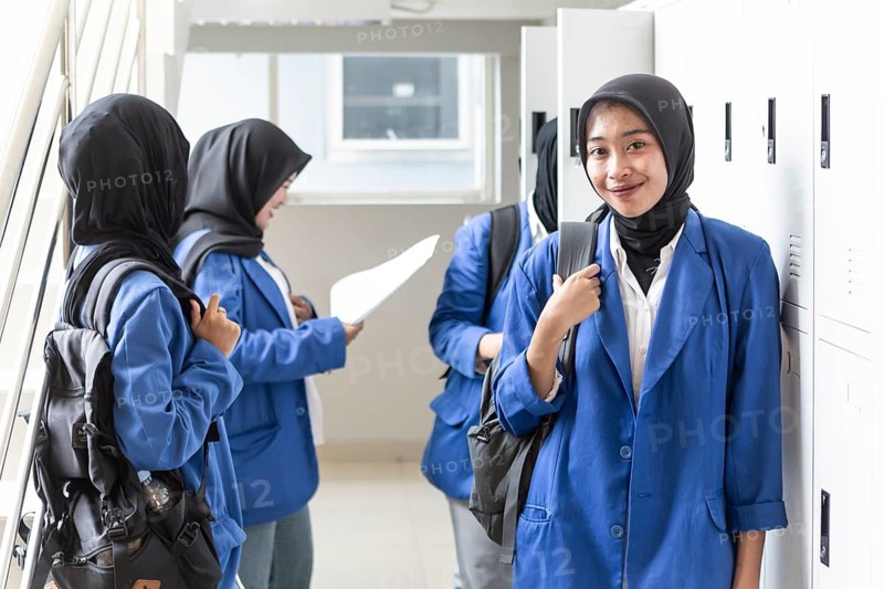 Asian Muslim students in blue blazers and hijabs stand and chat in a school corridor. They carry backpacks and documents, ready to start a new academic year with enthusiasm